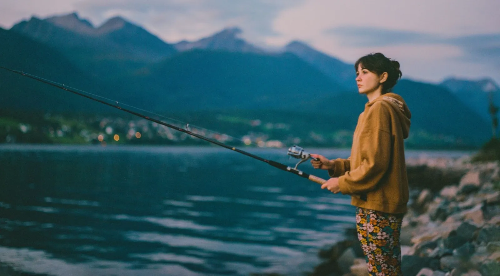A young angler casting from a rocky shore at twilight beside a calm fjord