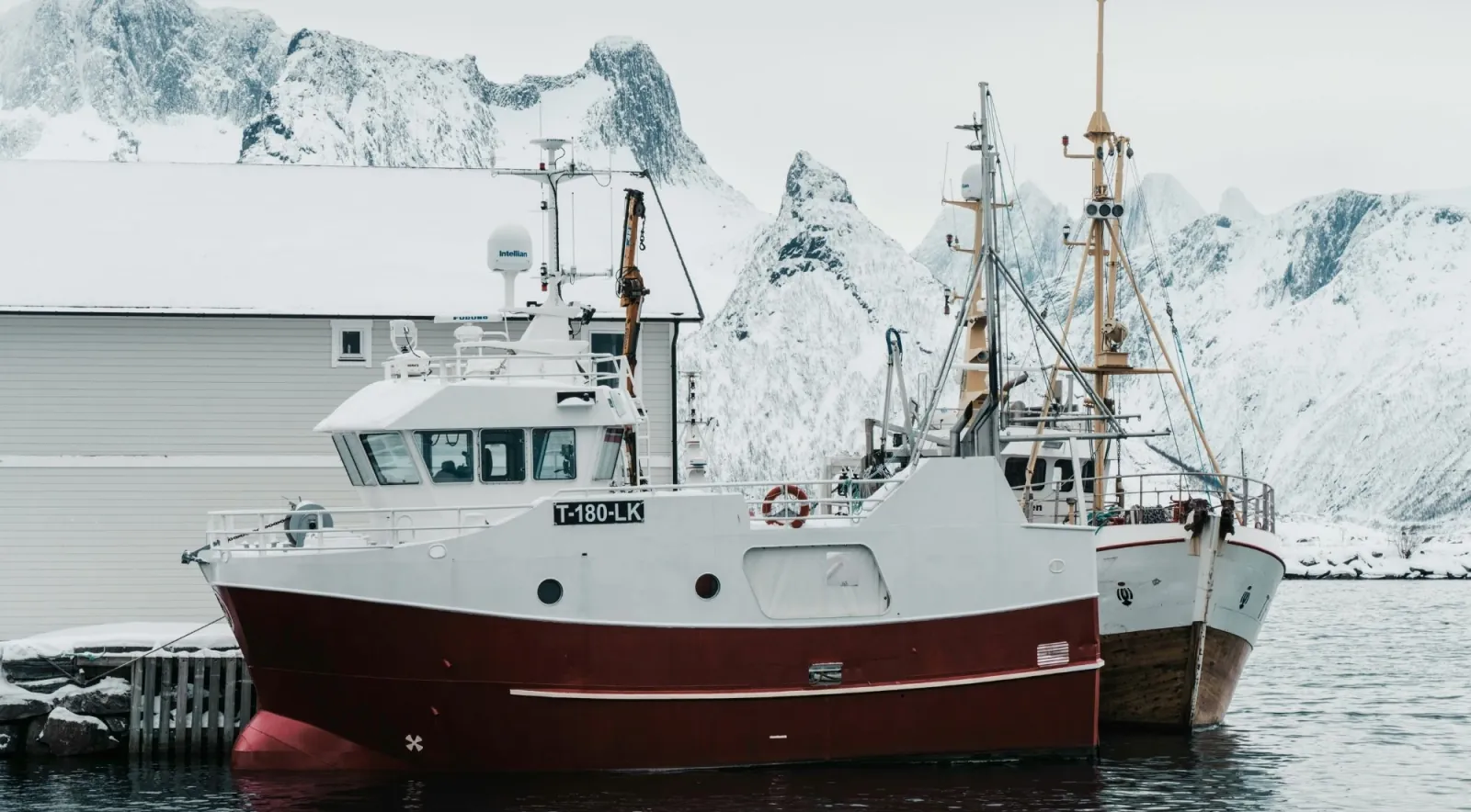 A red and white fishing trawler moored in a snowy harbour below jagged peaks